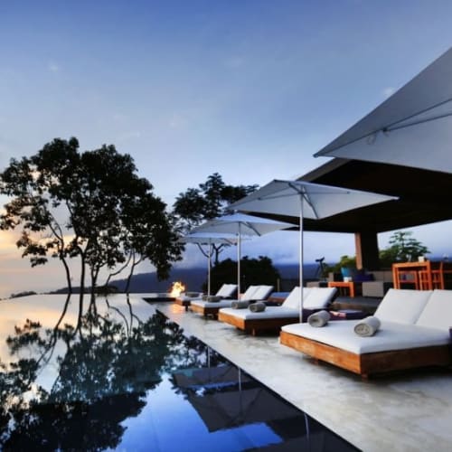 Luxury hotel outdoor area with infinity pool, lounge chairs, and umbrellas overlooking the ocean during sunset.