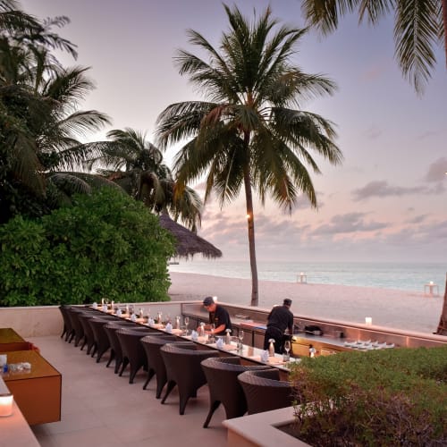 Outdoor dining area with a long table and chairs set up near the beach at sunset, with two staff members preparing the table under palm trees.