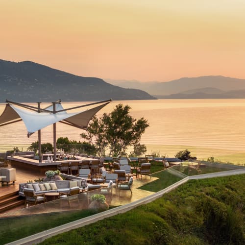 Outdoor terrace restaurant with modern seating and a large white sail shade overlooking a calm sea and distant mountains at sunset.