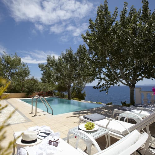 A luxurious hotel terrace with a pool, surrounded by trees and overlooking the sea, with sun loungers and flowers.