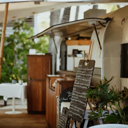 Außenbereich eines Cafés mit Holzmöbeln und einem geöffneten Fenster eines Metall-Imbisswagens, umgeben von Grünpflanzen und weißen Tischen mit Stühlen.