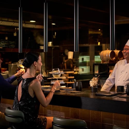 Guests enjoying drinks at the bar while a chef smiles behind the counter in a restaurant.