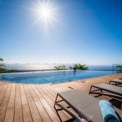 Sonnenbeschienener Infinity-Pool mit Meerblick, Holzboden und Liegestühlen mit blauen Handtüchern unter klarem blauem Himmel.