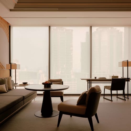 Moderne Hotellzimmer mit großen Fenstern, die einen Blick auf die Stadt bieten, eingerichtet mit einem Sofa, Sesseln und Schreibtischen.