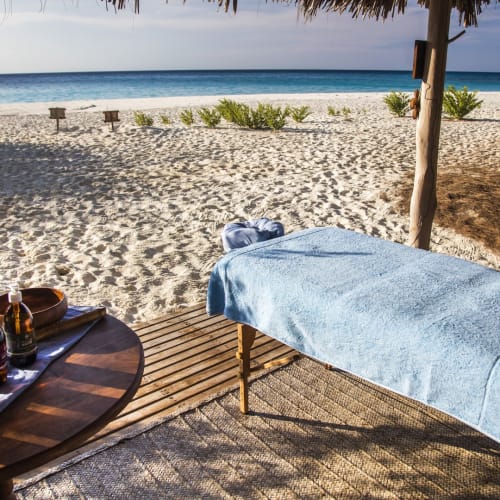 Eine Massageliege, die mit einem blauen Handtuch bedeckt ist, auf einer Holzterrasse mit Blick auf einen Sandstrand und den Ozean mit verstreuten Büschen.