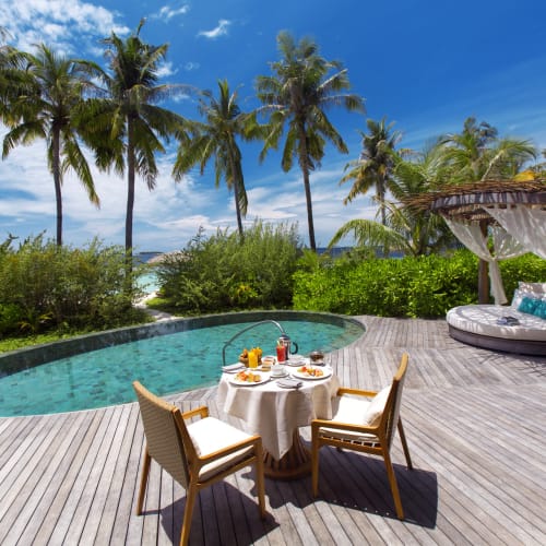 A luxurious outdoor setting on Milaidhoo Island in the Maldives featuring a private pool, a dining table with chairs, and a shaded lounge area amidst palm trees.