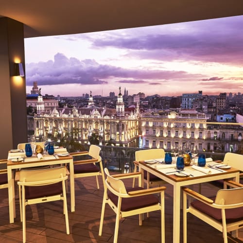 Outdoor restaurant with tables and chairs on a terrace overlooking a cityscape at sunset.