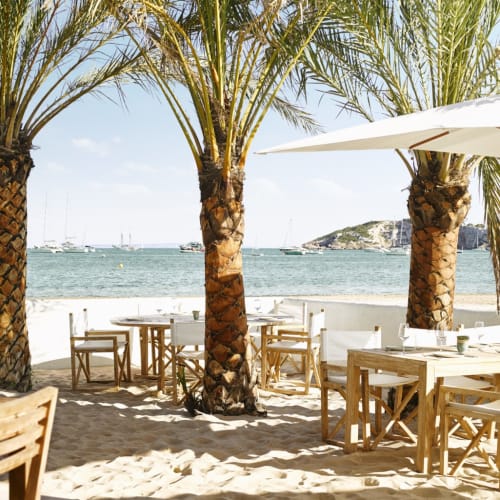 A chic beachside restaurant terrace at Nobu Hotel Ibiza, with wooden tables and chairs, large umbrellas, and palm trees overlooking the sea.