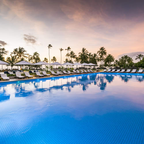 Ein luxuriöser Poolbereich mit Liegestühlen und Sonnenschirmen, umrundet von Palmen bei Sonnenuntergang.