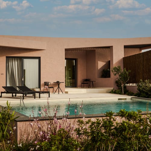 Modernes einstöckiges Gebäude mit Flachdach, beiger Fassade, einem Schwimmbecken vorne, zwei schwarzen Liegestühlen und einem kleinen Tisch mit Stühlen auf der Terrasse, umgeben von Grün und Blumen unter einem teils bewölkten Himmel.