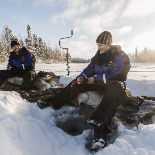 Two men dressed in winter clothing are ice fishing on a frozen lake surrounded by snow-covered trees in Finland, with an ice auger nearby and a bright sky above.