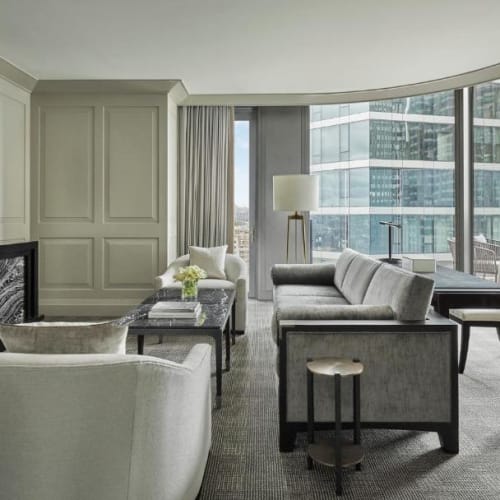 Ein modernes Hotelzimmer mit großem Fenster, das Wolkenkratzer in der Stadt zeigt, ausgestattet mit Sofas, Sesseln, einem Kamin und einem Flachbildfernseher.