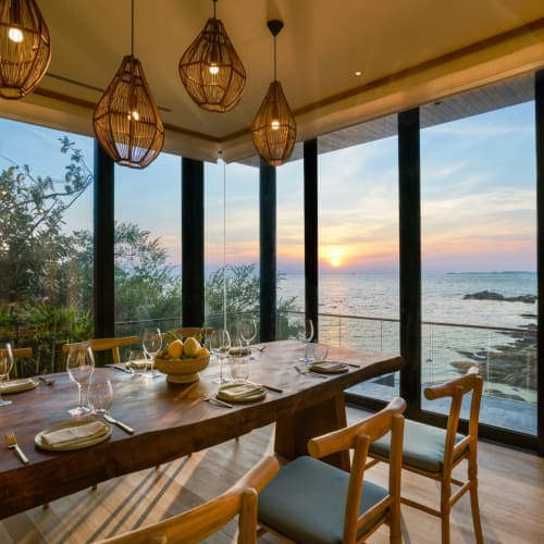 Elegantes Esszimmer mit einem großen Holztisch, gedeckt mit Glaswaren und Tellern, mit Blick auf den Strand und Sonnenuntergang über dem Meer durch bodentiefe Fenster.
