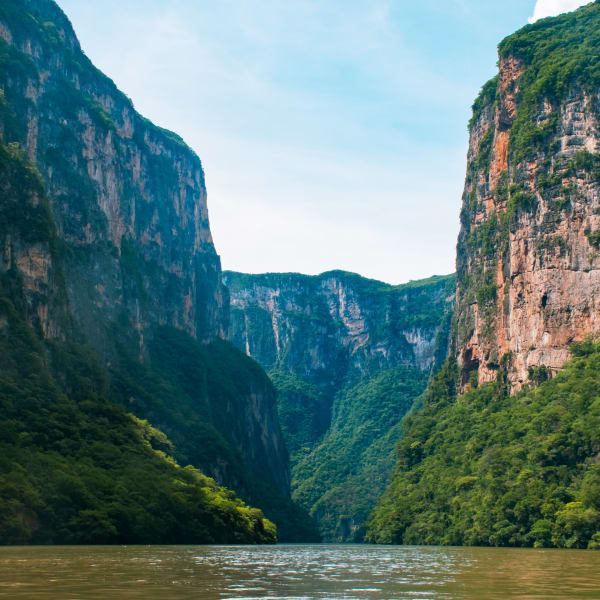 Fluss fließt durch eine tiefe Schlucht mit steilen felsigen Klippen, die mit Grün bedeckt sind, unter einem teilweise bewölkten blauen Himmel.