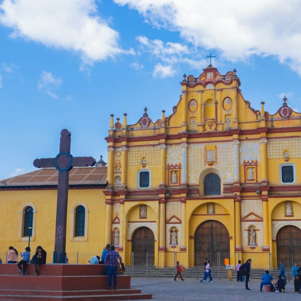 Helle gelbe koloniale Kirchenfassade in San Cristóbal de las Casas mit einem großen Kreuzdenkmal im Vordergrund und Menschen, die über einen sonnigen Platz gehen.