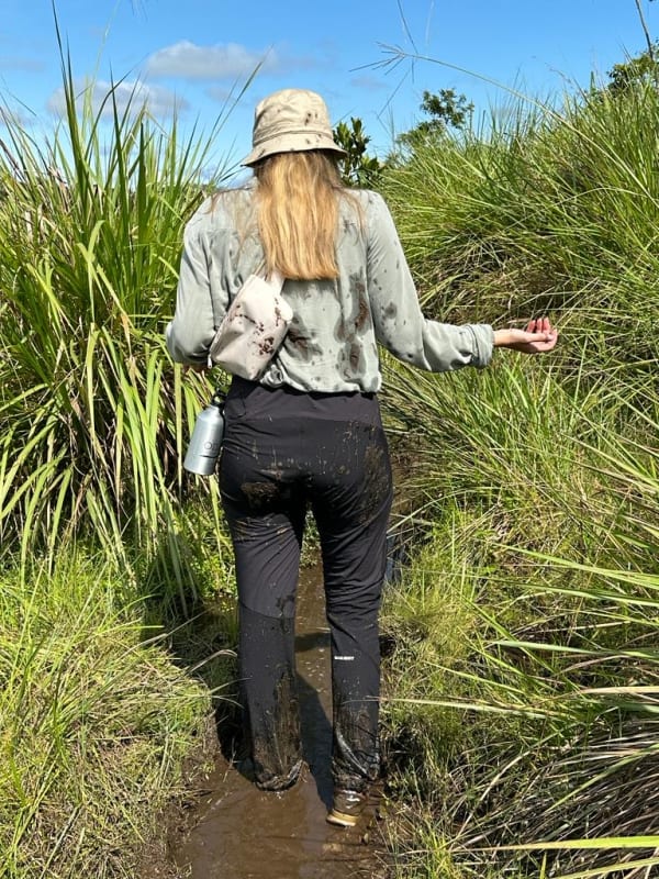 Eine Frau läuft durch einen matschigen, grasbewachsenen Weg in einer natürlichen Umgebung, gekleidet mit Outdoor-Ausrüstung und Hut.