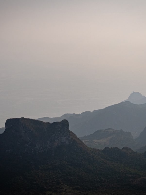 Mist-covered mountain range with multiple ridges fading into the distance over the Yemen coastline under a pale sky with two birds flying.