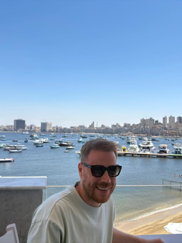 A man wearing sunglasses smiling while sitting at an outdoor table with a waterfront view of numerous boats and a city skyline in the background under a clear blue sky.