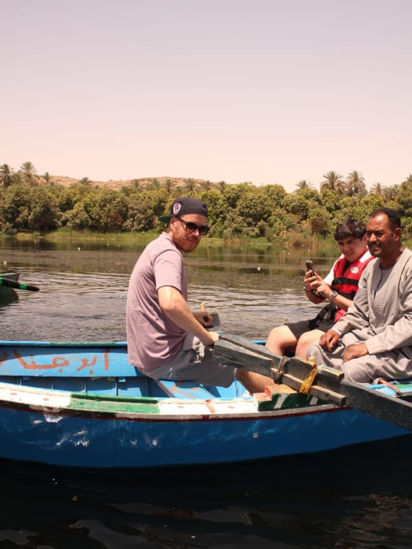 Three men sitting in a blue wooden boat on a calm river with green vegetation and palm trees along the riverbank under a clear sky.