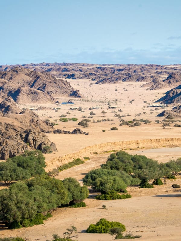 A semi-arid landscape with scattered green trees, surrounded by rugged mountains under a partly cloudy sky.