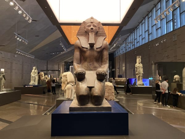 Große steinerne Statue eines altägyptischen Pharaos kniend im Grand Egyptian Museum in Kairo, umgeben von weiteren ägyptischen Artefakten und Besuchern.