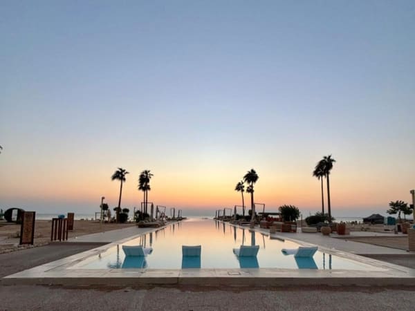 Langer rechteckiger Pool mit weißen Liegestühlen am Rand, beidseitig von Palmen gesäumt, bei klarem Sonnenuntergang in einem Strandresort.