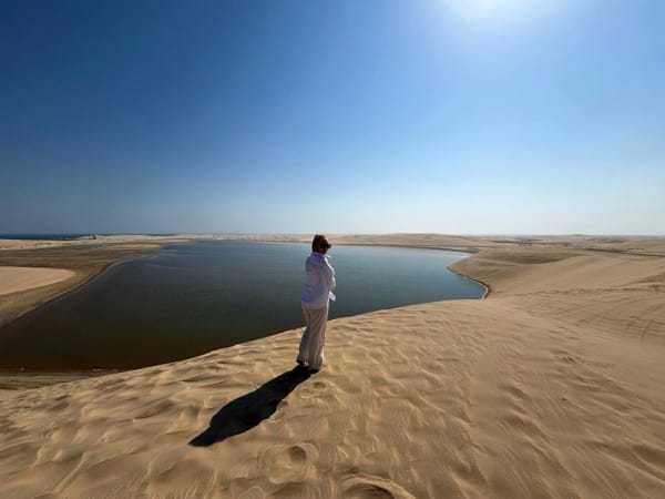 Eine Person steht auf sandigen Dünen und blickt auf ein ruhiges Binnenmeer unter klarem blauem Himmel in Katar.