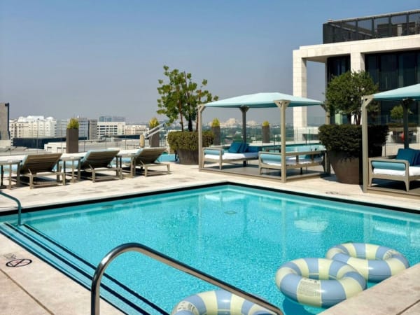 Ein Dachpool im Mandarin Oriental Doha, umgeben von Liegestühlen, Pavillons mit blauen Kissen, großen Topfpflanzen und Stadthäusern im Hintergrund bei klarem Himmel.
