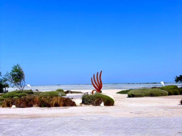 Sandige Landschaft mit grünen Büschen und einer roten dekorativen Skulptur in Form einer Hand mit ausgestreckten Fingern unter klarem blauem Himmel im Zulal Wellness Resort bei Chiva-Som.