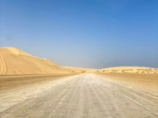 Eine gerade, staubige Straße, die durch eine Wüstenlandschaft mit großen Sanddünen unter klarem blauem Himmel führt.