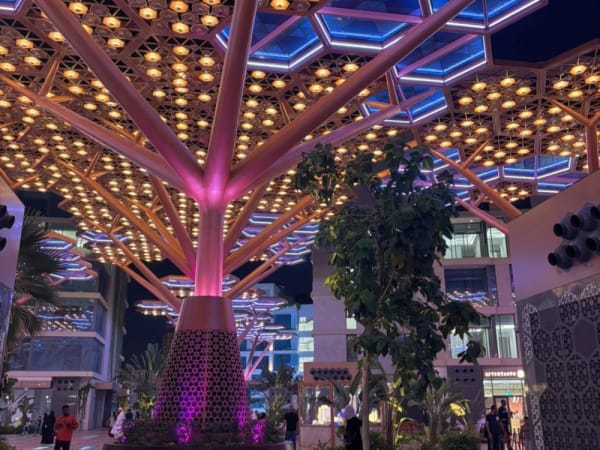 Nachtansicht von Gewan Island Crystal Walk mit großen baumähnlichen Strukturen, beleuchteten sechseckigen Überdachungen und umgebenden modernen Gebäuden mit spazierenden Menschen und Grünanlagen.