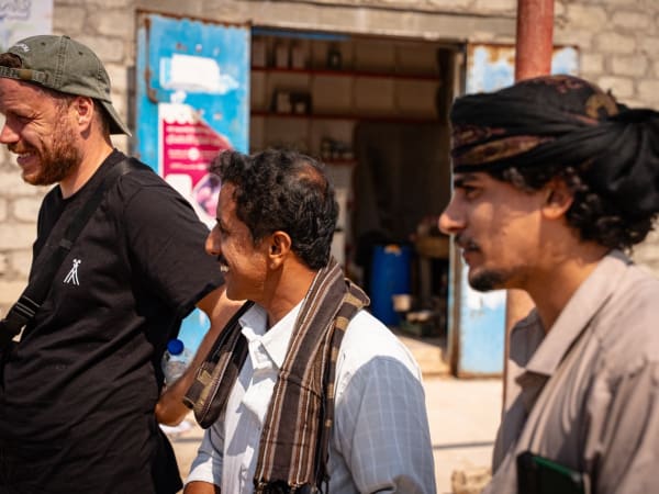Three men standing outdoors in front of a rustic stone building with open blue doors, engaged in conversation and smiling.