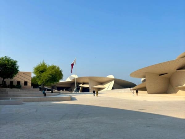 Weitläufiger Innenhof mit modernen, skulpturalen Gebäuden unter klarem blauem Himmel im Nationalmuseum in Doha, mit einer Flaggenstange und der Flagge von Katar sowie einigen Spaziergängern.