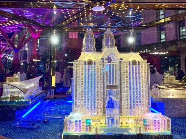 Ein detailliertes beleuchtetes Miniaturmodell von The Pearl, Doha, in einem Glaskasten mit lila und blauem Umgebungslicht und Spiegelungen von Personen im Hintergrund.