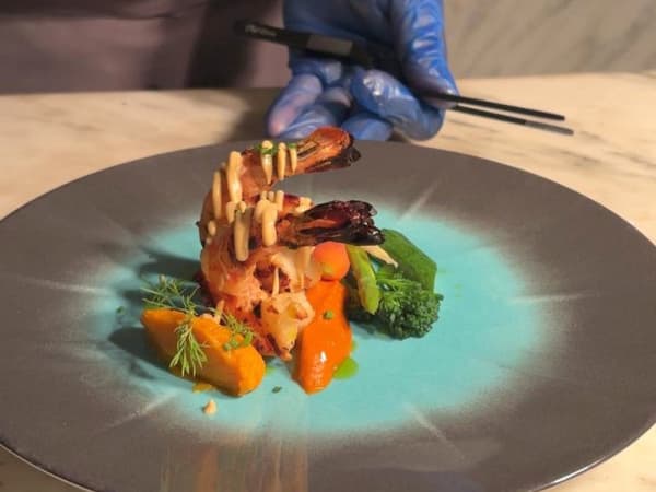 Ein angerichteter Teller mit gegrillten Garnelen, die mit Soße beträufelt sind, begleitet von Gemüse wie Brokkoli und Karottenpüree, serviert auf einem blau-grauen Teller mit einer blau behandschuhten Hand, die im Hintergrund Essstäbchen hält.