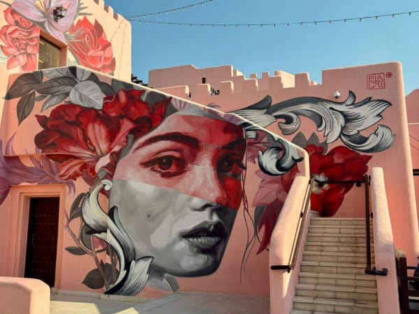 Ein großes Wandgemälde zeigt das Gesicht einer Frau mit Blumen- und Ornamentaldesigns auf den Wänden eines rosa Gebäudes neben einer Treppe unter klarem blauem Himmel.