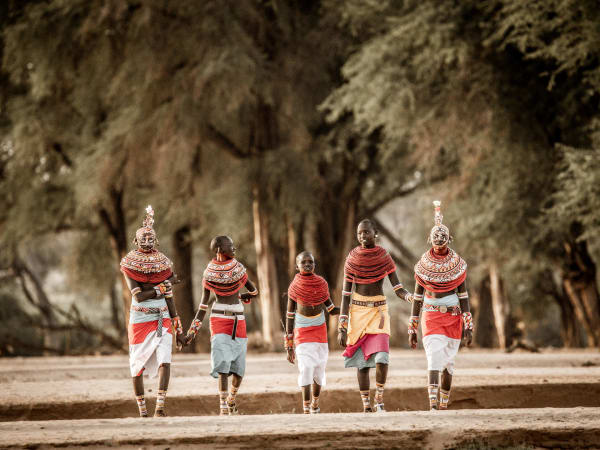 Six members of a local Samburu tribe walking in traditional attire on dirt ground with trees in the background.