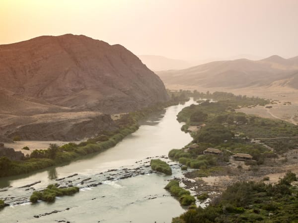 Ein Fluss, der durch eine bergige Wüstenlandschaft mit grüner Vegetation entlang seiner Ufer in Namibia fließt.