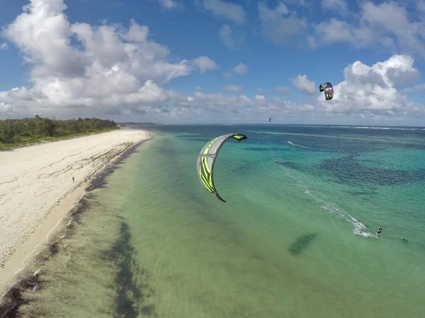 Kitesurfer genießen den Wind und die Wellen an einem tropischen Strand mit klarem Wasser und wolkigem Himmel.
