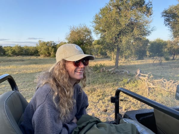 A woman with long hair wearing a hat and sunglasses, smiling while sitting in a vehicle in a grassy, tree-filled landscape.