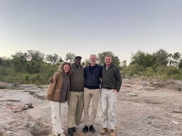 Four people standing closely together outdoors with trees and rocks in the background.