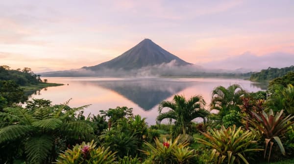 Ein ruhiger Blick auf den Vulkan Arenal, der sich im ruhigen Wasser eines Sees bei Sonnenaufgang spiegelt, umgeben von dichter tropischer Vegetation in Costa Rica.