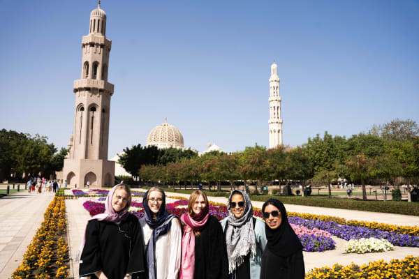 Eine Gruppe von fünf Frauen mit Kopftüchern steht vor bunten Blumenbeeten an der Sultan-Qaboos-Grand-Moschee in Oman unter klarem blauem Himmel.