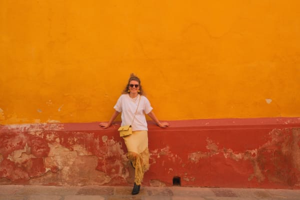 Eine Frau mit Sonnenbrille, weißem T-Shirt und gelbem Rock lehnt an einer strukturierten roten und orangen Wand im Freien.