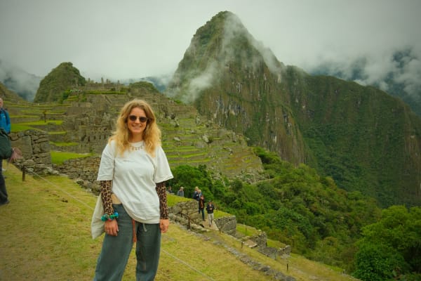 Eine Frau mit Sonnenbrille steht auf grasbewachsenen Terrassen bei den Ruinen von Machu Picchu, im Hintergrund ein teilweise von Nebel bedeckter Berg.