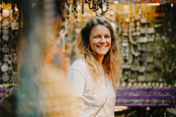 A woman with long, wavy hair smiling in front of a colorful, jewelry-filled background.