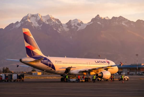 Ein LATAM Airlines Flugzeug steht bei Sonnenuntergang auf dem Rollfeld eines Flughafens mit schneebedeckten Berggipfeln im Hintergrund.