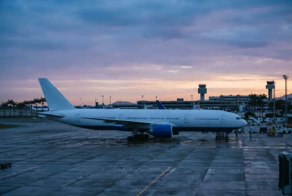 Ein großes weißes Verkehrsflugzeug steht auf einem Flughafen-Vorfeld bei der Morgendämmerung oder Abenddämmerung mit einem Terminalgebäude und Kontrolltürmen im Hintergrund.