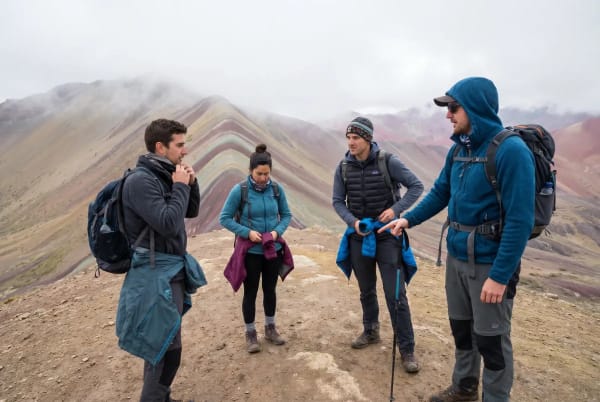 Vier Wanderer in Outdoor-Bekleidung stehen auf einem felsigen Pfad vor bunten, gestreiften Bergkämmen und wolkigem Himmel, vermutlich am Rainbow Mountain in Peru.