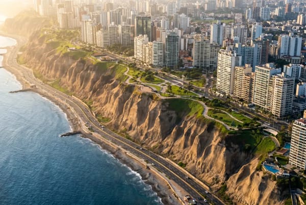 Luftaufnahme von Miraflores, Lima, mit Küstenklippen, einer Straße entlang der Küste, hohen Gebäuden und einer urbanen Skyline im Hintergrund bei Sonnenuntergang.